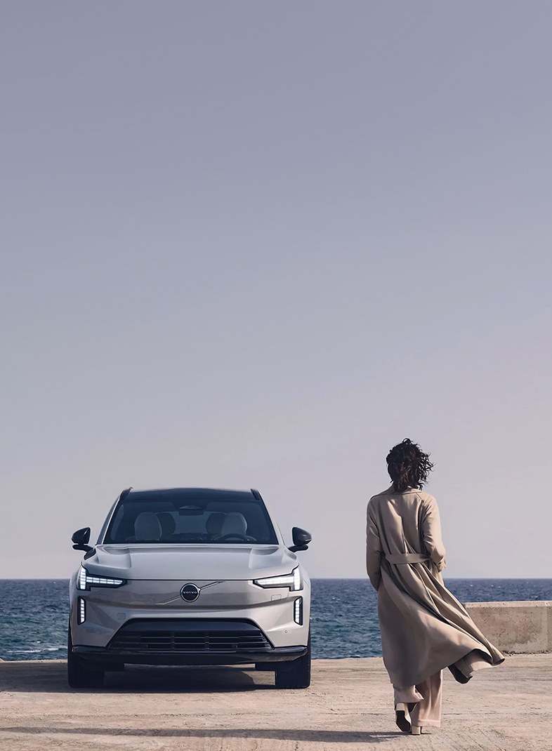 Volvo EX90 estacionado à beira-mar, com uma mulher caminhando ao lado